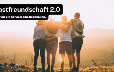 Vier Personen stehen auf einem Hügel und umarmen sich, während sie in den Sonnenuntergang blicken. Der Text „Gastfreundschaft 2.0“ und „Mehr als ein Service: eine Begegnung.“ ist oben links im Bild zu sehen.